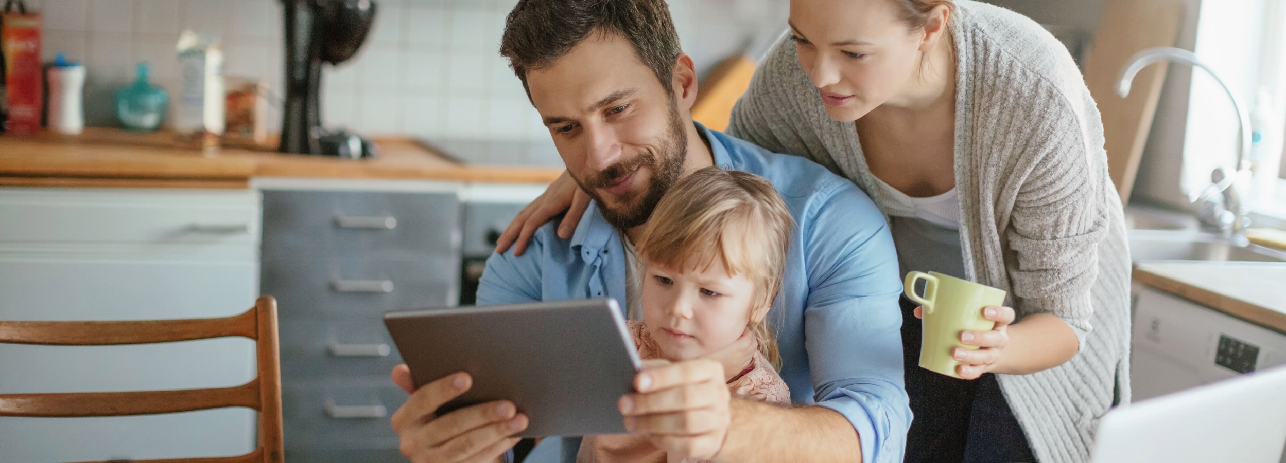Ein junger Vater sitzt mit einem Kleinkind auf dem Schoß in einer Küche. Seine Frau beugt sich von hinten über seine Schulter, um einen Blick auf das Tablet zu werfen, dass der Vater in der Hand hält.