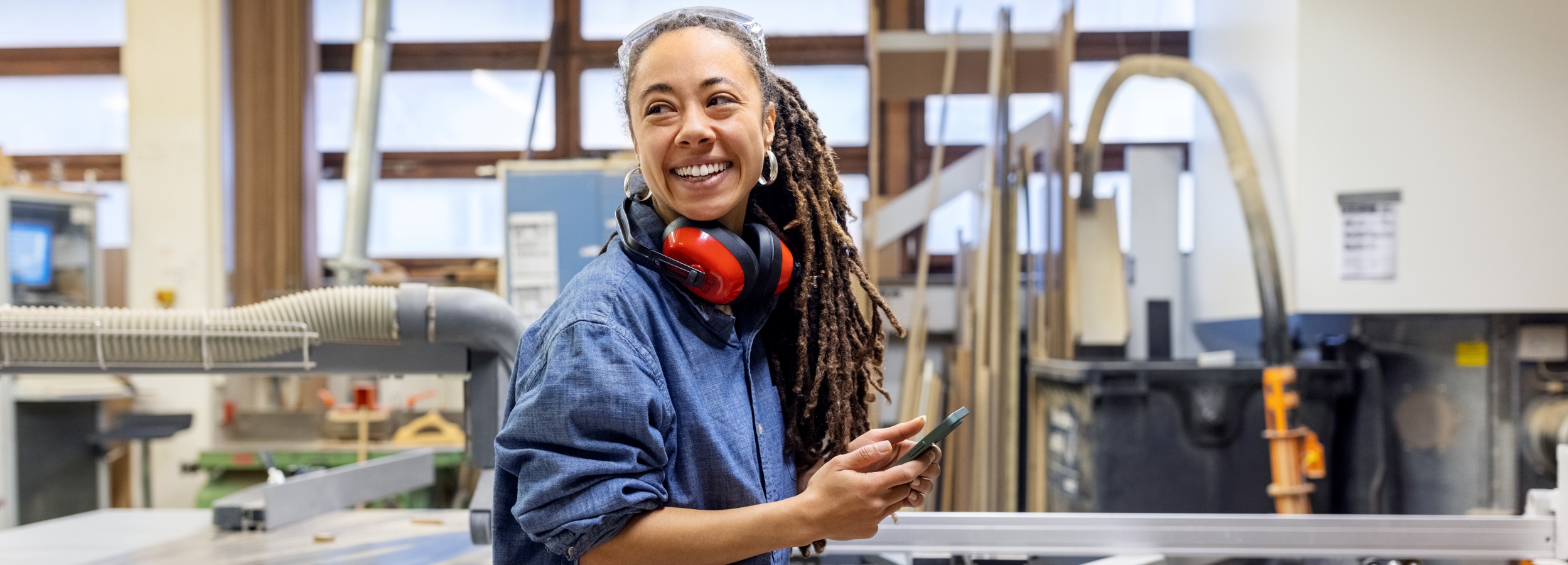 Das Bild zeigt eine lächelnde Frau in einer Werkstatt, die ihr Smartphone hält und sich wahrscheinlich über ihre geleistete Arbeit freut.  Im Detail ist eine Frau mittleren Alters mit Dreadlocks und einem strahlenden Lächeln zu sehen. Sie trägt eine blaue Arbeitsbluse, eine Schutzbrille auf dem Kopf und rote Gehörschützer, die um ihren Hals hängen. In ihren Händen hält sie ein Smartphone.  Der Hintergrund zeigt eine industrielle Werkstattumgebung mit Holzbearbeitungsmaschinen, Holzbrettern und Staubabsauganlagen. Die Werkstatt ist hell erleuchtet, wahrscheinlich durch Tageslicht, das durch die Fenster fällt.  Die Atmosphäre des Bildes ist positiv und professionell. Die lächelnde Frau strahlt Selbstvertrauen und Zufriedenheit aus, was darauf hindeutet, dass sie ihre Arbeit gerne macht und stolz auf ihre Fähigkeiten ist. Die Werkstatt wirkt sauber und organisiert, was einen professionellen und sicheren Arbeitsplatz suggeriert.