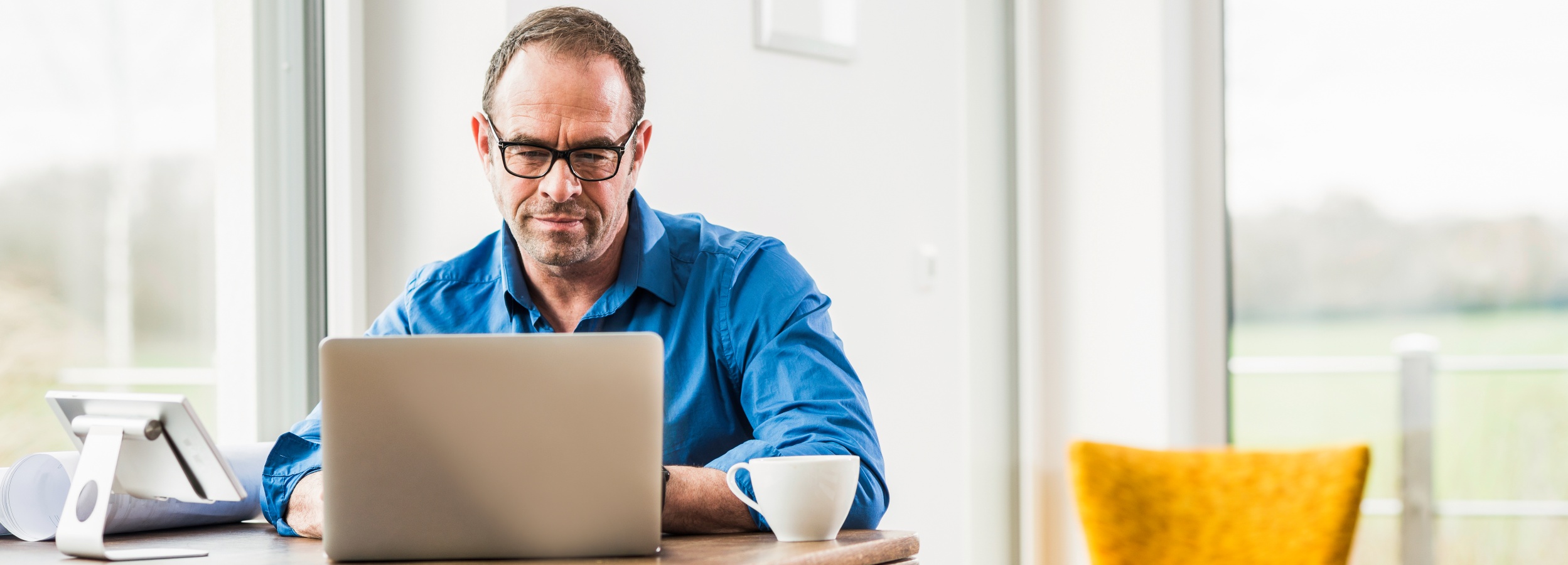 Ein Mann mittleren Alters sitzt an einem Holztisch und arbeitet konzentriert an einem Laptop, während er eine Tasse Kaffee daneben hat, die eine häusliche Szene zeigt.  Die Komposition ist eine Innenaufnahme, die eine Person in einer Arbeitsumgebung zeigt. Der Mann sitzt vor einem Laptop, mit dem Blick auf den Bildschirm gerichtet. Links vom Laptop befindet sich ein Tablet mit einem Ständer und einige Papierrollen, die möglicherweise Arbeitsunterlagen darstellen. Auf der rechten Seite steht eine weiße Kaffeetasse. Das Bild ist horizontal ausgerichtet und erstreckt sich über einen größeren Bereich, was den Eindruck von Raum vermittelt. Die Szene suggeriert einen Moment der Konzentration und Arbeit.