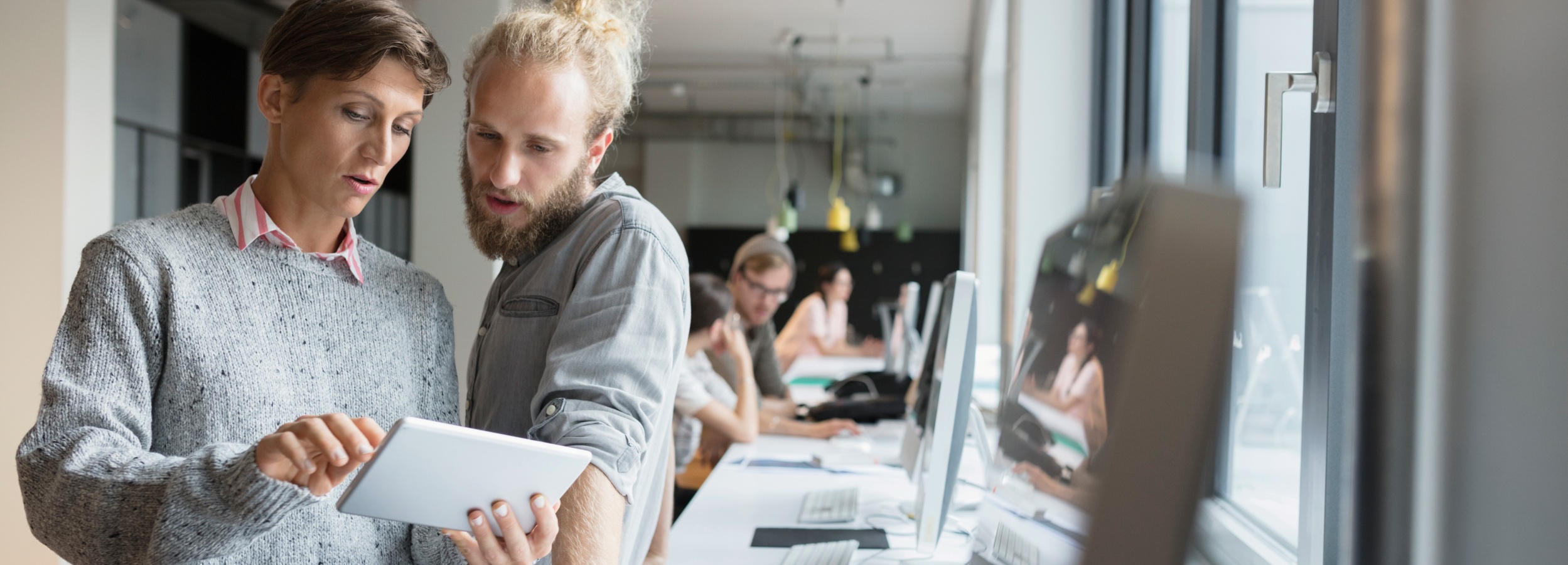 Zwei Kollegen besprechen konzentriert ein digitales Dokument auf einem Tablet in einem modernen Büro.  Auf dem Bild sind zwei Personen im Vordergrund zu sehen, die offensichtlich zusammenarbeiten. Die Person links, deren Oberkörper und Kopf zu sehen sind, hat kurze, dunkle Haare und trägt einen grauen Strickpullover über einem gestreiften Hemdkragen. Ihr Blick ist nach unten auf das Tablet gerichtet, das sie mit beiden Händen hält. Ihre Zeigefinger berühren den Bildschirm, als ob sie etwas erklären oder auswählen würden. Ihr Gesichtsausdruck ist konzentriert und ernst.  Neben ihr steht eine Person, von der ebenfalls der Oberkörper und Kopf zu sehen sind. Diese Person hat blondes Haar, das zu einem Dutt hochgebunden ist, und einen Bart. Sie trägt ein hellgraues Hemd. Ihr Kopf ist leicht zur Seite geneigt, und sie blickt ebenfalls auf das Tablet, ihr Mund ist leicht geöffnet, was auf eine engagierte Diskussion oder eine Frage hindeutet.  Im Hintergrund ist ein Großraumbüro zu erkennen. Mehrere Arbeitsplätze mit Computern sind in einer Reihe aufgestellt. Durch das Fenster dringt helles Tageslicht, das die Szene beleuchtet. An der Decke hängen moderne Lampen in verschiedenen Farben. Weitere Personen sind im Hintergrund zu sehen, die an ihren Computern arbeiten, jedoch unscharf dargestellt. Die Atmosphäre ist geschäftig und doch ruhig, geprägt von Konzentration und Teamarbeit. Die Farbpalette ist überwiegend neutral mit Grau-, Weiß- und Beigetönen, was dem Raum eine moderne und professionelle Ausstrahlung verleiht. Es gibt keine Tiere auf dem Bild. Die Stimmung ist professionell, konzentriert und kollaborativ.