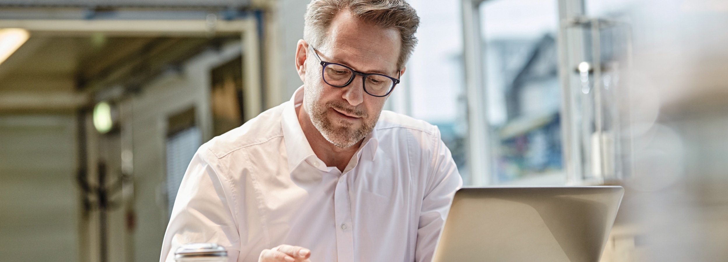 Ein Mann mittleren Alters mit Brille und einem weißen Hemd arbeitet konzentriert an seinem Laptop in einem hellen, modernen Büro.  Das Bild zeigt einen Mann mittleren Alters, der offensichtlich fokussiert ist, während er auf einen Laptop schaut, der im Vordergrund seiner rechten Seite platziert ist. Er trägt eine schlichte weiße Hemdbluse, die leicht zerknittert ist, was auf eine aktive Arbeitsphase hindeutet. Seine Haare sind kurz geschnitten und leicht grau meliert, ebenso wie sein Bartansatz. Er trägt eine moderne Brille mit dunklem Rahmen. Sein Blick ist nach unten gerichtet, auf den Bildschirm, wobei seine rechte Hand leicht erhoben ist, als würde er gerade eine Eingabe tätigen oder etwas auf dem Bildschirm zeigen. Im linken Vordergrund ist ein Teil eines silbernen, zylindrischen Objekts zu sehen, möglicherweise eine Kaffeetasse oder ein Behälter.  Die Umgebung scheint ein Büro zu sein, angedeutet durch die unscharfen Hintergründe, die industrielle oder moderne Architekturmerkmale aufweisen könnten, wie Metallträger und eine große Fensterfront, durch die Tageslicht einfällt. Das Licht ist weich und diffus, was zu einer ruhigen und nachdenklichen Atmosphäre beiträgt. Die Stimmung ist professionell und konzentriert, ohne gehetzt zu wirken. Es ist ein Moment der stillen Arbeit und des Nachdenkens dargestellt, in dem der Mann tief in seine Aufgabe vertieft ist.