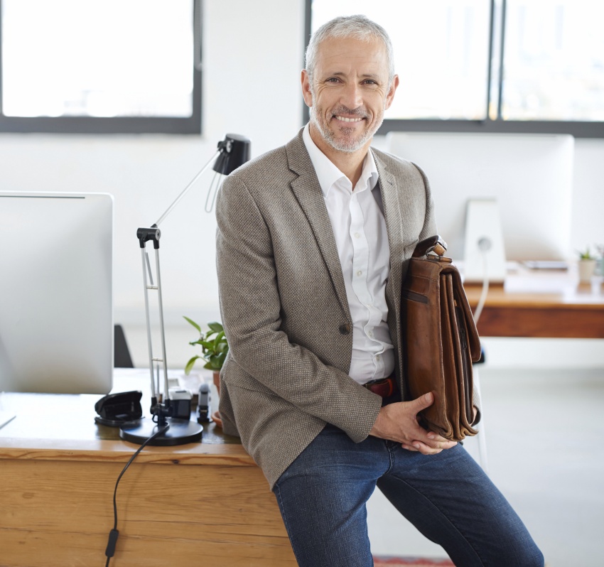Ein älterer Mann mit kurzen grauen Haaren sitzt an einem Schreibtisch in einem hellen Büro. Er trägt ein weißes Hemd und ein graubraunes Sakko und lächelt in die Kamera. Neben ihm steht ein brauner Aktenkoffer. Auf dem Schreibtisch sind ein Computerbildschirm und eine Schreibtischlampe zu sehen. Im Hintergrund befinden sich große Fenster und ein Regal.