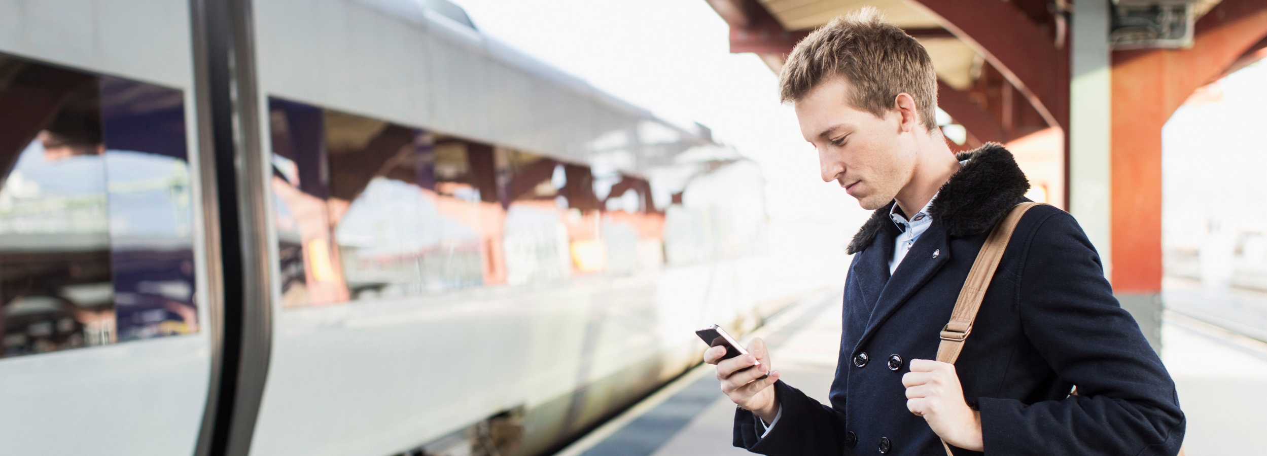 Ein junger Mann steht auf einem Bahnsteig und schaut auf sein Handy, während ein Zug daneben hält. Auf dem Bild ist ein junger Mann mit hellbraunem Haar zu sehen, der einen dunklen Mantel mit einem auffälligen Kragen aus Kunstpelz trägt. Unter dem Mantel trägt er ein hellblaues Hemd mit feinem Muster. Er hat eine Umhängetasche aus beigefarbenem Leder über der Schulter. Der Mann steht in einer leichten Seitenansicht und blickt konzentriert auf sein Smartphone, das er in der rechten Hand hält. Sein Gesichtsausdruck ist nachdenklich. Im Hintergrund ist ein langer, moderner Zug zu sehen, der an einem Bahnsteig hält. Die Türen des Zuges sind geschlossen und die Fenster spiegeln die Umgebung wider, was eine leicht verschwommene Optik erzeugt. Über dem Bahnsteig befindet sich eine Überdachung, deren roten Trägerkonstruktion im sanften Licht zu erkennen ist. Die Atmosphäre des Bildes ist ruhig und urban, mit einem Hauch von Vorfreude oder Erwartung, die mit Reisen verbunden ist. Das helle, diffuse Licht deutet auf einen bewölkten Tag oder eine leichte Dämmerung hin. Die Stimmung ist gelassen und fokussiert, wobei der Mann im Mittelpunkt des Geschehens steht, obwohl er in seine digitale Welt vertieft ist.