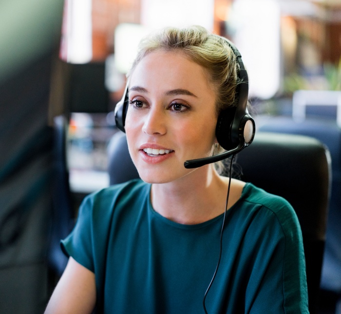 Das Bild zeigt eine freundliche Call-Center-Mitarbeiterin, die mit einem Kunden spricht.  Im Vordergrund des Bildes ist eine junge Frau mit blonden Haaren zu sehen, die mit einem Headset mit Mikrofon ausgestattet ist und ein dunkelgrünes Oberteil trägt. Sie sitzt an einem Schreibtisch in einem Büro, und der Hintergrund ist unscharf, es sind jedoch Computerbildschirme und andere Büromöbel erkennbar. Das Licht fällt auf ihr Gesicht und hebt ihre freundlichen Gesichtszüge hervor; ihre Augen sind leicht nach oben gerichtet, und sie lächelt leicht, was auf ein positives Gespräch mit einem Kunden hindeutet. Die Atmosphäre ist professionnell und aufmerksam, und die Stimmung ist ruhig und konzentriert.