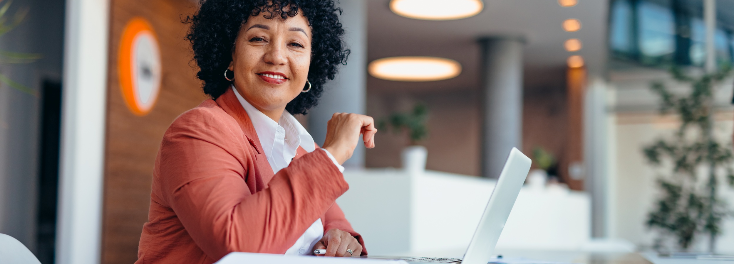 Die Aufnahme zeigt eine lächelnde Geschäftsfrau, die in einem modernen Büro sitzt und in die Kamera blickt.  Im Vordergrund des Bildes sitzt eine Frau mittleren Alters mit dunklem, lockigem Haar und einem warmen Lächeln. Sie trägt einen weißen Kragen eines Hemdes und darüber einen korallenfarbenen Blazer. Große silberne Ohrringe zieren ihre Ohren, und ein zarter Ring ist an ihrer linken Hand zu sehen. Ihr Blick ist direkt auf den Betrachter gerichtet, was eine einladende und selbstbewusste Ausstrahlung erzeugt. Ihre rechte Hand ruht leicht auf ihrem Schoß, während ihre linke Hand etwas nach oben gestreckt ist, was auf eine entspannte, aber aufmerksame Haltung hindeutet. Vor ihr liegt ein aufgeklappter Laptop auf einem Tisch.  Der Hintergrund ist unscharf und deutet auf eine moderne Büroumgebung hin. Man erkennt helle, ovale Deckenleuchten, die für eine angenehme Beleuchtung sorgen. Graue Säulen und Holzakzente in der Wandgestaltung deuten auf ein stilvolles und professionelles Umfeld hin. Im Hintergrund ist auch eine grüne Pflanze zu erkennen, die einen Hauch von Natur in den Raum bringt. Die allgemeine Atmosphäre ist hell, professionell und positiv. Die Stimmung ist optimistisch und selbstsicher, vermittelt durch das Lächeln der Frau und die moderne, aufgeräumte Umgebung.