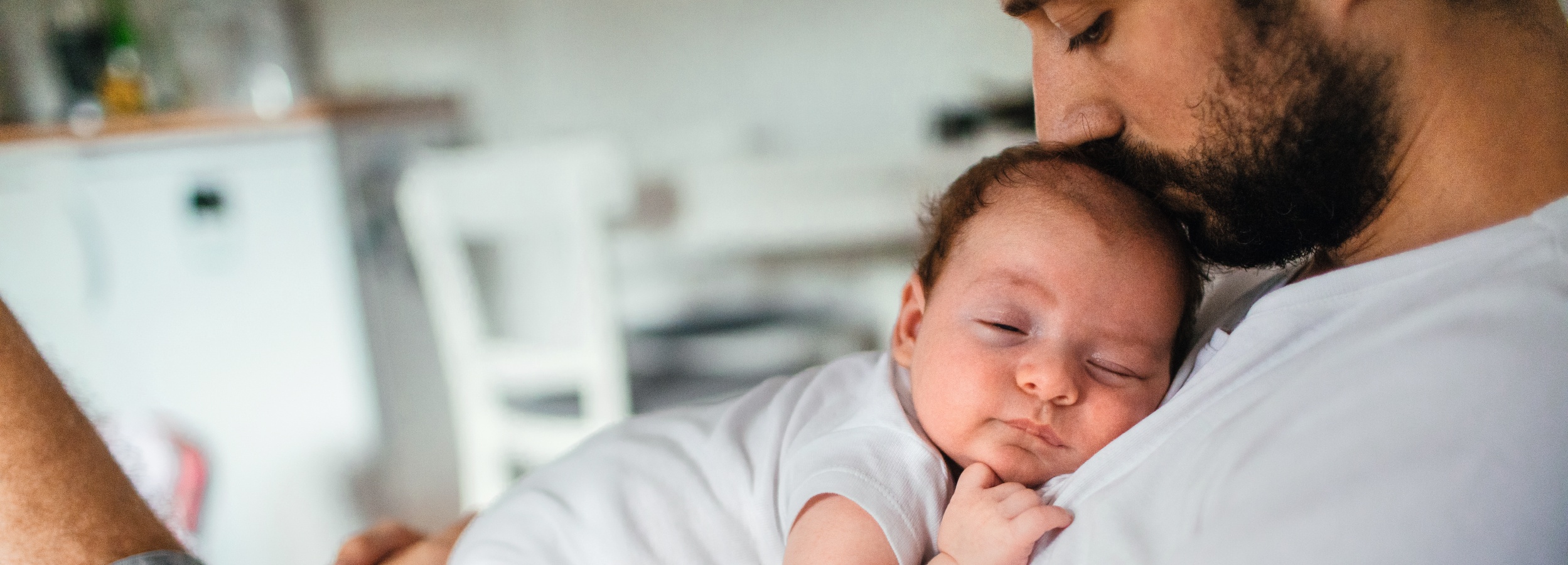 Dieses Bild zeigt die zärtliche Umarmung eines Vaters und seines schlafenden Babys.  Das Bild ist eine Nahaufnahme, die sich auf die intime Bindung zwischen einem Vater und seinem Neugeborenen konzentriert. Der Vater, dessen Gesicht teilweise verdeckt ist, beugt sich über das Baby und küsst sanft dessen Stirn. Er hat einen Bart und trägt ein weißes T-Shirt. Das Baby, das ebenfalls ein weißes Outfit trägt, schläft friedlich in den Armen des Vaters. Sein Gesicht ist dem Betrachter zugewandt, und es liegt eine tiefe Ruhe auf seinen Zügen. Die winzige Hand des Babys ruht auf seiner Brust. Der Hintergrund ist unscharf und deutet auf eine häusliche Umgebung hin, möglicherweise eine Küche oder ein Wohnzimmer, mit verschwommenen Formen von Möbeln, die aber nicht weiter von Bedeutung sind, da der Fokus klar auf der Vater-Kind-Beziehung liegt.  Die Atmosphäre des Bildes ist geprägt von Wärme, Liebe und Geborgenheit. Die sanfte Beleuchtung und die Nahaufnahme verstärken das Gefühl der Intimität und des Schutzes. Die Bildkomposition und die weichen Konturen tragen zu einer friedlichen und beruhigenden Stimmung bei, die die tiefe emotionale Verbindung und die bedingungslose Liebe zwischen Eltern und Kind hervorhebt. Es vermittelt ein Gefühl der Ruhe und des Vertrauens, das typisch für die frühen Momente des Elternseins ist.