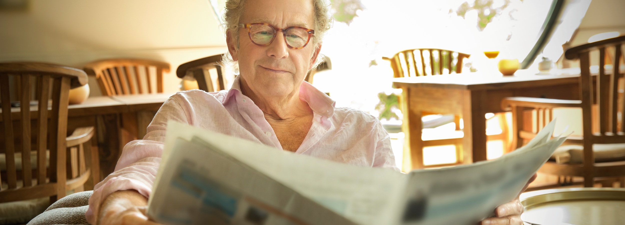 Ein älterer Herr liest entspannt eine Zeitung in einem hellen Raum.  Auf dem Bild ist ein älterer Mann mit grauem Haar und einer Brille mit Schildpattrahmen zu sehen, der auf einem Sessel sitzt und eine Zeitung liest. Er trägt ein hellrosa Hemd mit offenen Knöpfen am Kragen. Seine Hände halten die Zeitung, die im Vordergrund unscharf ist. Der Hintergrund ist ebenfalls unscharf und zeigt eine Inneneinrichtung mit Holztischen und Stühlen, die in warmem Sonnenlicht gebadet sind. Auf einem der Tische steht ein Glas mit einer gelben Flüssigkeit. Die Atmosphäre des Bildes ist ruhig, gelassen und friedlich. Das natürliche Licht und die warmen Farben erzeugen eine einladende und behagliche Stimmung, die an einen entspannten Morgen oder Nachmittag erinnert. Der Mann strahlt Zufriedenheit und Gelassenheit aus, während er sich seiner Lektüre widmet.